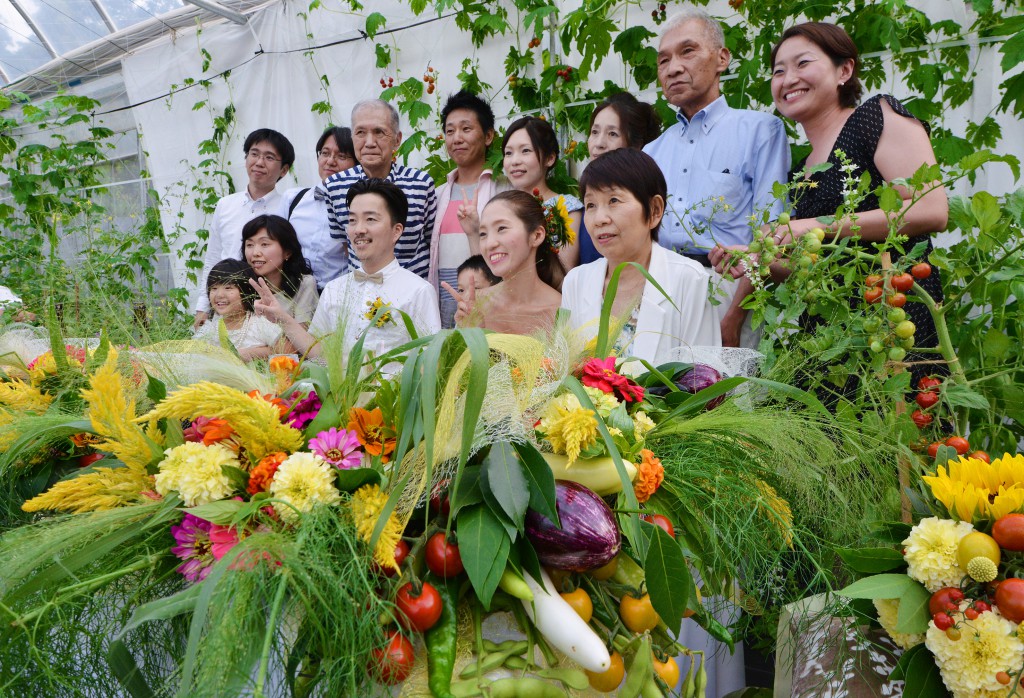 Happy pair (center) taking pictures with parents and relatives at head table decorated with flowers and vegetables. The arrangement uses more than 10 kinds of colorful vegetables including egg plants, tomatoes and green soybeans in pods.  
